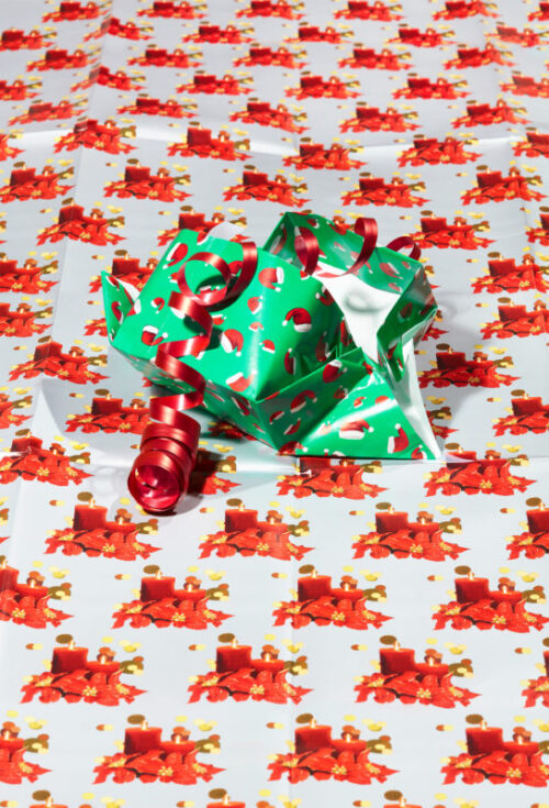 Litt brukt gavepapir/julepapir i grønt med rødt mønster og rødt gavebånd, lagt oppå lyst julepapir med stearinlysmønster i rødt
