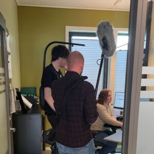 Kameramann og lydmann står inne på et kontor sammen med medarbeider, som sitter på stol foran PC