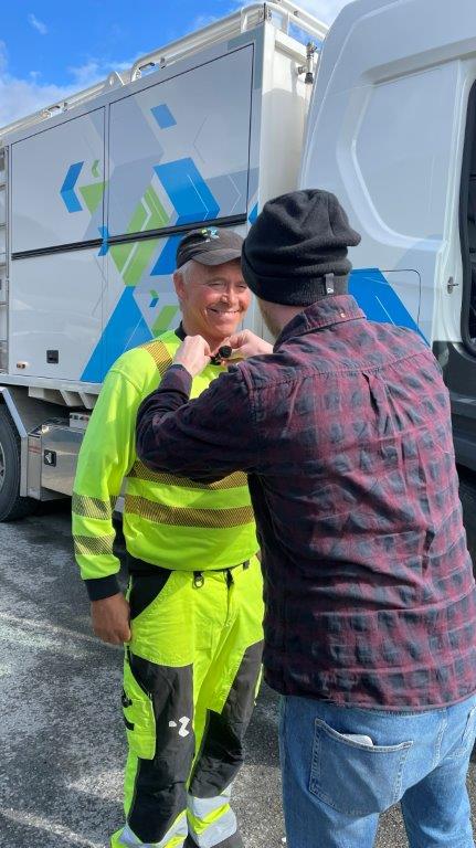 Sjåfør Torje i gult arbeidstøy, smiler mens en mann fester en mikrofon på ham